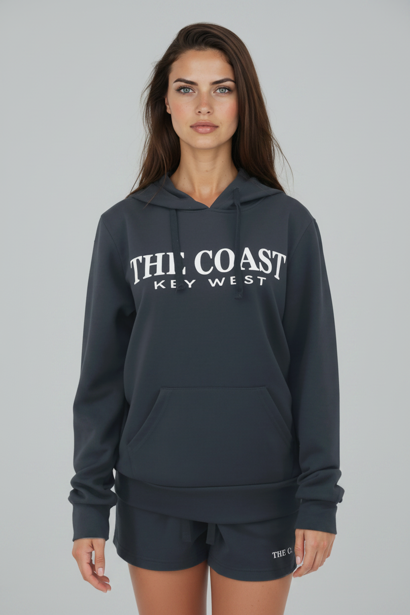 A woman modeling a dark gray hoodie with 'THI' COAST KEY WEST' printed in white across the chest, paired with matching dark gray shorts against a solid gray studio background.