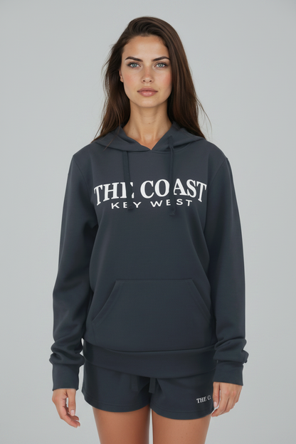 A woman modeling a dark gray hoodie with 'THI' COAST KEY WEST' printed in white across the chest, paired with matching dark gray shorts against a solid gray studio background.
