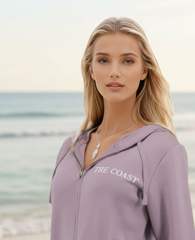 A blonde woman wearing a lavender zip-up hoodie with 'THE COAST' printed in white on the chest, standing against a blurred beach background.
