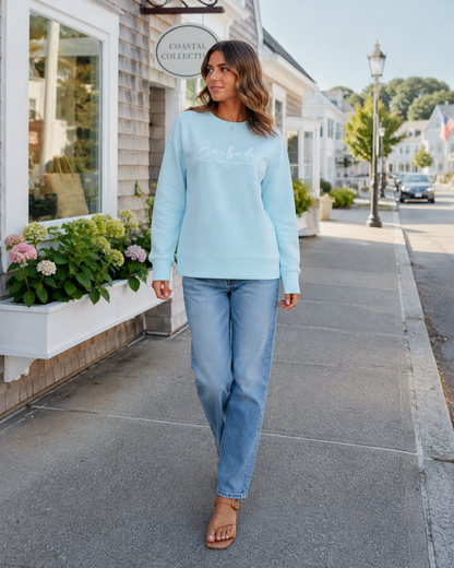 A woman walking on a sunny sidewalk wearing a light blue long-sleeve crewneck sweatshirt with 'Coastal Collective' printed in white script across the chest, paired with light-wash blue jeans and tan slide sandals.