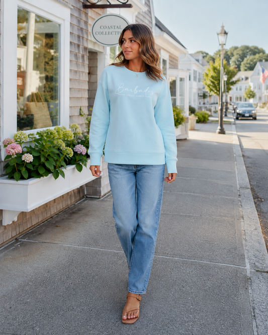 A woman walking on a sunny sidewalk wearing a light blue long-sleeve crewneck sweatshirt with 'Coastal Collective' printed in white script across the chest, paired with light-wash blue jeans and tan slide sandals.