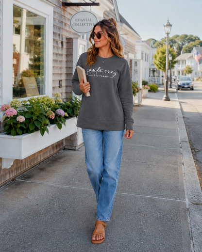 A woman walking on a sunny town street wearing a dark gray crewneck sweatshirt with 'sacred craft' printed in white script across the chest, paired with light blue jeans and brown leather sandals.