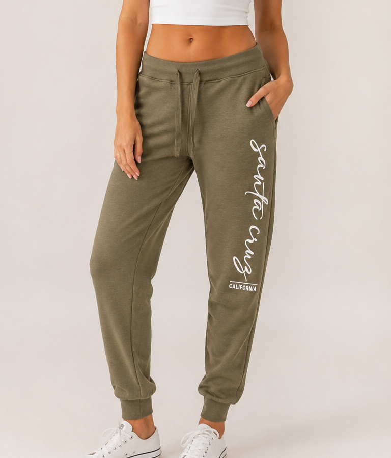 A person wearing olive green jogger sweatpants with a white script graphic reading 'Santa Cruz' and 'CALIFORNIA' on the lower leg, paired with a white crop top and white sneakers, against a light gray studio background.
