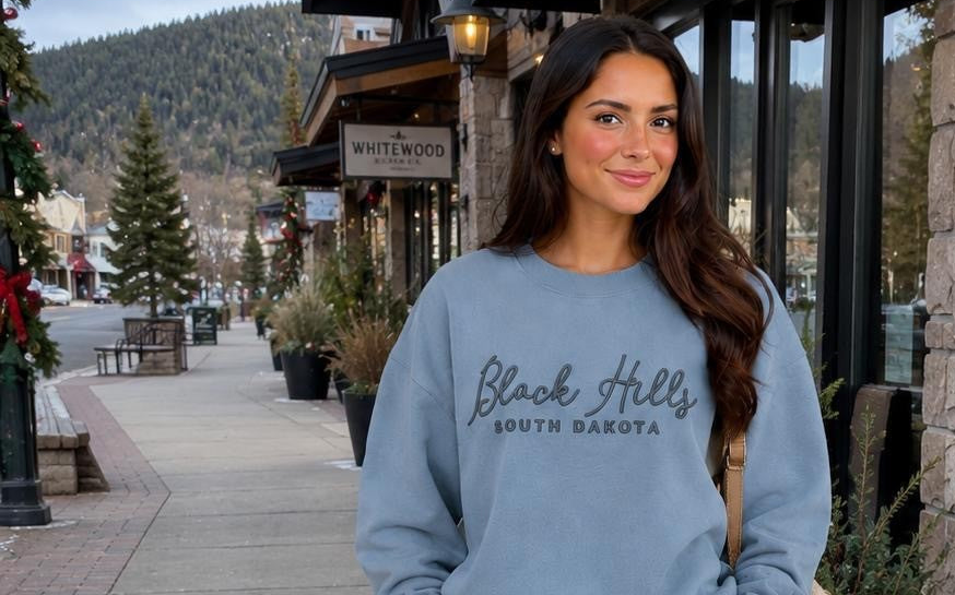 A woman stands on a town street wearing a light blue crewneck sweatshirt with 'Black Hills South Dakota' printed in gray across the chest. The background shows storefronts, evergreen trees, and mountains.