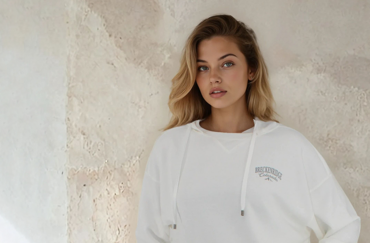 A woman with blonde hair wearing a white pullover hoodie with a dark gray graphic on the left chest, standing against a textured, light beige plaster wall.