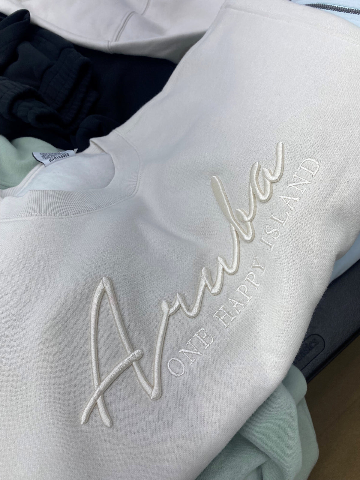 A close-up of a white crewneck sweatshirt featuring 3D embroidered cursive text reading 'Anuba' and the phrase 'ONE HAPPY ISLAND' in matching thread, layered over a pile of other folded garments.