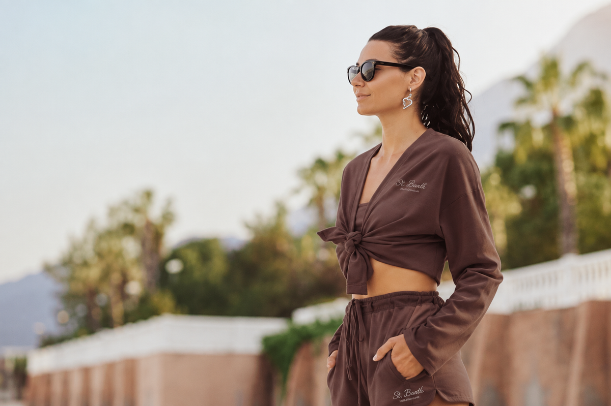 A woman wearing a matching brown lounge set with a long-sleeve tie-front crop top and drawstring pants, styled with dark sunglasses and earrings, standing outdoors against a blurred background of palm trees and mountains.